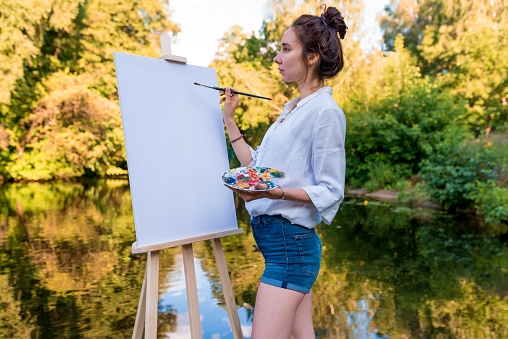 beautiful girl artist, summer park by lake, river pond, draws picture, palette with paints, brush white canvas painting, easel stand. White shirt, denim shorts. Green forest background water