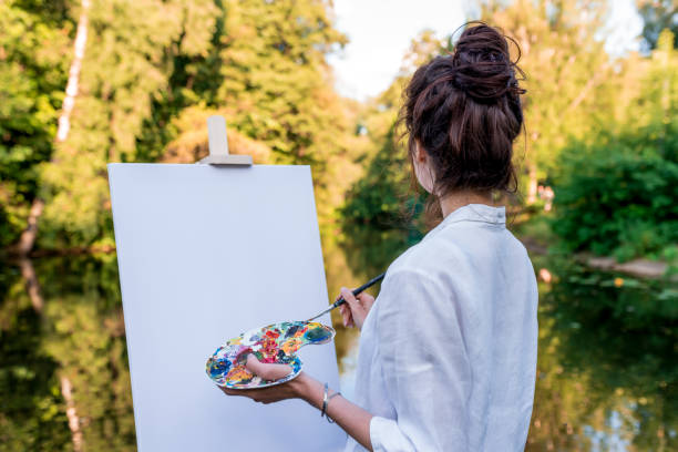 Girl artist, in summer park lake, view from back, river pond, draws picture, palette with paints, brush white canvas painting, easel stand. White shirt. Green forest background with trees and water. stock photo