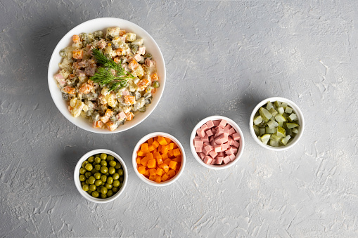 Traditional Russian Olivier salad in a white bowl and main ingredients green peas, boiled carrots, ham, pickles on a gray background. Top view, copy-space, no people