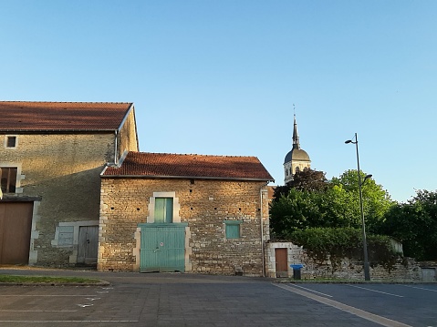 Rustic picture Frenche village Andelot with ancient houses, barn with colourful green or blue doors and brickwall. 19th century. Trees and ancient small church in the background.