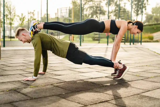 Fit caucasian couple in a sportwear standing in the pose of the bridge on each other, doing yoga on a sportsground