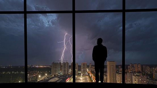 The man standing near the panoramic window on the night lightning background
