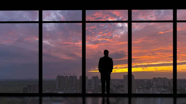 Photo of The man standing near the panoramic window on the city sunset background
