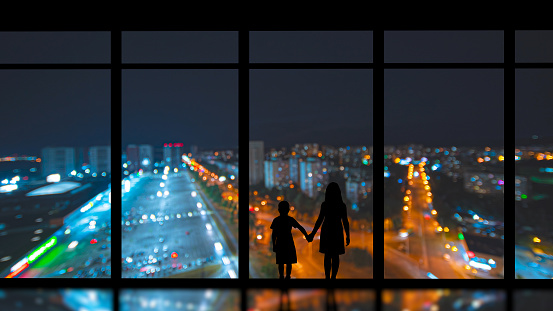 The girl and boy standing near the panoramic window against the evening city