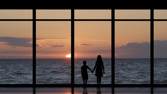 The kids standing near the panoramic window against the sea sunset
