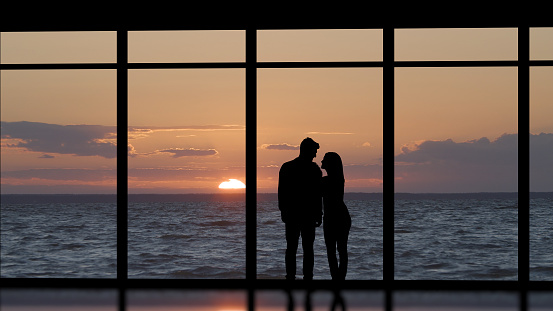 The man and woman standing near the panoramic window against the sea sunset