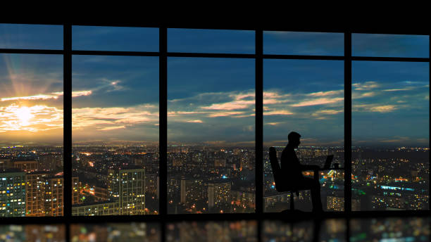 l’homme d’affaires s’asseyant près de la fenêtre panoramique contre le lever du soleil de ville - lighting equipment office building night city photos et images de collection