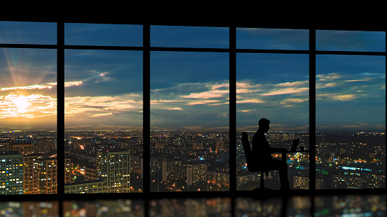 The businessman sitting near the panoramic window against the city sunrise