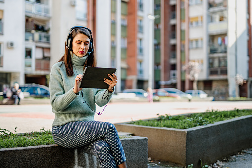 Young smiling woman an adult university student using a digital tablet and headphones, listening to a class online outdoors in the park