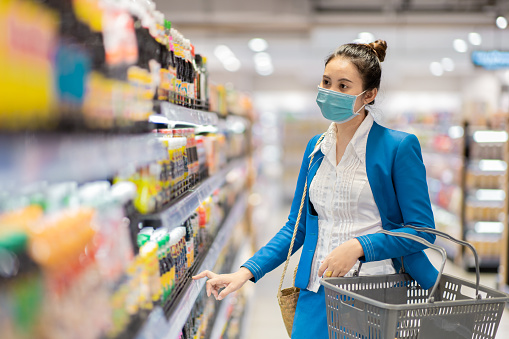 asian young businesswoman who is wearing face mask and walking in supermarket aisle to buy products with trolley