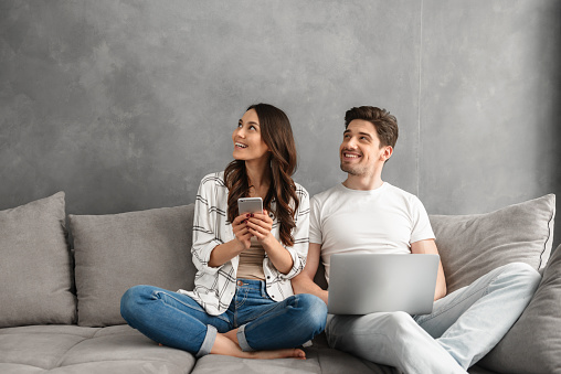 Beautiful man and woman sitting together on couch in gray interior and looking aside on copyspace while using laptop and smartphone