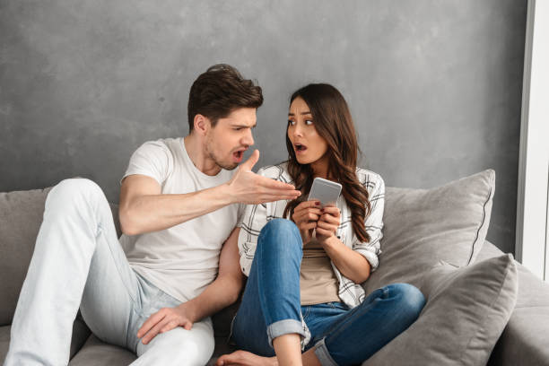 집에서 소파에 함께 앉아 있는 불쾌한 부부는 남자가 손에 스마트 폰을 들고 있는 여성에게 소리를 지르며 흰색 배경 위에 고립되어 있습니다. - attentively 뉴스 사진 이미지