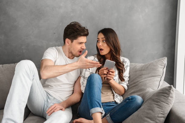 displeased couple sitting together on sofa at home while man shouting on woman who holding smartphone in hand, isolated over white background - attentively imagens e fotografias de stock