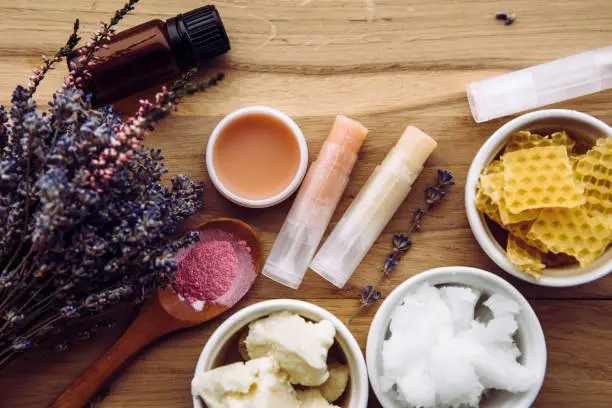 Photo of Ingredients for homemade lip balm: shea butter, essential oil, mineral color powder, beeswax, coconut oil. Homemade lip balm lipstick mixture with ingredients scattered around.