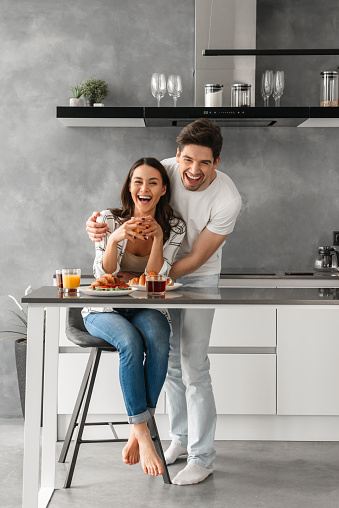 Full length portait of cheerful couple smiling and looking on you while having dinner in modern apartment