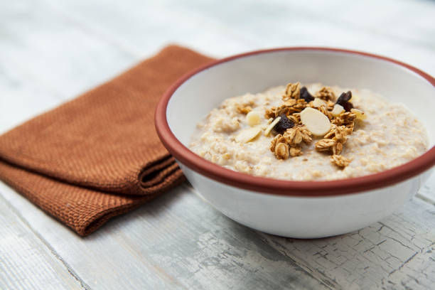 porridge mit granola in einer weißen schale mit braunem trimm gekrönt, und braune serviette auf einem grob bemalten holztisch - oatmeal porridge oat raisin stock-fotos und bilder