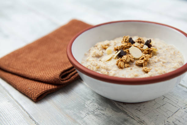 porridge condito con muesli in una ciotola bianca con finiture marroni e tovagliolo marrone su un tavolo di legno dipinto grezzo - oatmeal raisin porridge nut foto e immagini stock