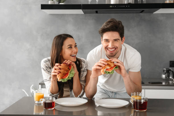 młoda i piękna para uśmiechnięta i jedząca kanapki na śniadanie w kuchni - propping zdjęcia i obrazy z banku zdjęć