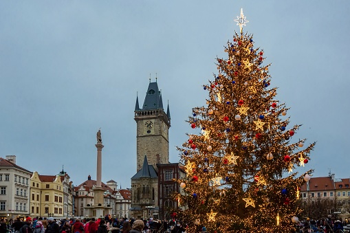 Prague, Czech Republic - December 12 2020: The town hall, Marian column and Christmas tree decorated with colorful lights and stars. Blue sky in the background. People in street.
