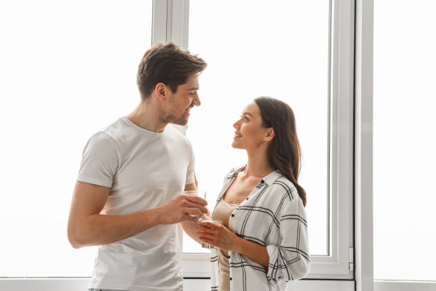 istock Married couple smiling, and looking at each other while standing in front of big window with glasses in hands 1292759382