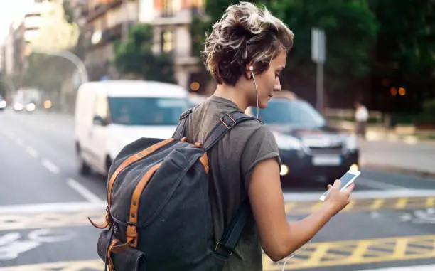 Photo of Student girl is usinf smartphone to find the address of the hostel. Young tomboy girl is listening to the music on her mobile phone while walking the street on a blurred urban background.