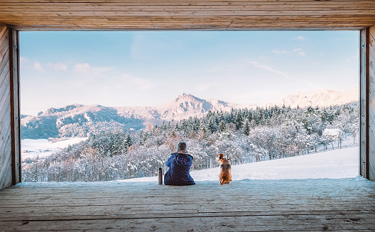 Woman with thermos flask sitting with her beagle dog in the big wooden hangar with a huge panoramic window and looking on the snowy mountain landscape. Human and pets relatives concept image.