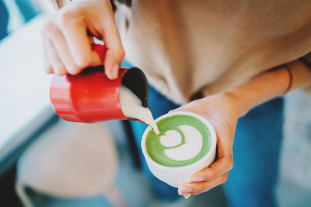 istock Barista making flower patterns while  preparing tasty and healthy matcha latte tea. Woman's hand pouring hot whipped milk into the cup with freshly made natural matcha tea in an asian coffee shop. 1292762083