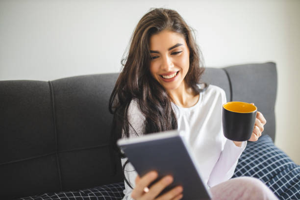 istock Beautiful young woman reading magazine on digital tablet and drinking coffee while sitting in bed at the morning 1292765664