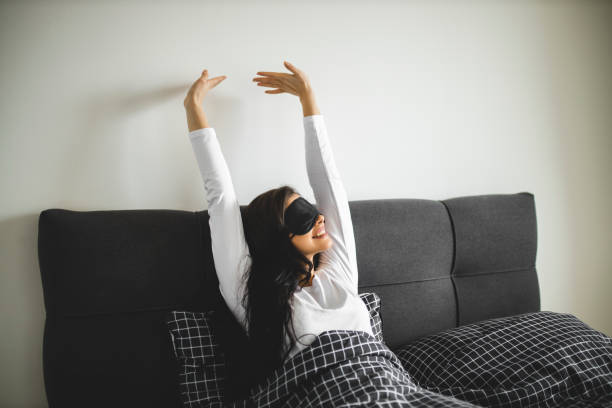 Young woman wearing sleeping mask and stretching while sitting in her bed Young woman wearing sleeping mask and stretching while sitting in her bed sleep eye mask stock pictures, royalty-free photos & images