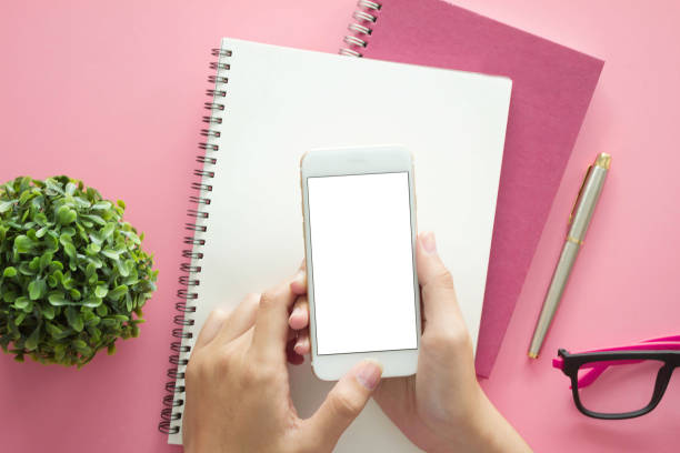 istock Mockup image of hands holding white mobile phone with blank white screen with  Modern pink office desk with laptop and other supplies for input the text on copy space Top view, flat lay. 1292769688