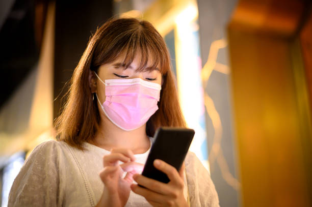 istock Young Asian woman with protective facemask using smart phone while walking through the downtown district. 1292773681