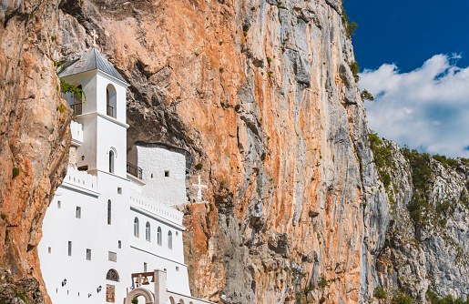 White Monastery Ostrog of the Serbian Orthodox Church situated against an almost vertical mountain with blue cloudy sky in background. Montenegro travel, Europe.