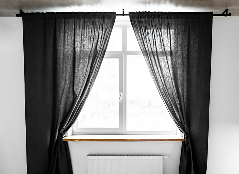 Panel pair cotton curtains tied back at the modern window. Two separate linen curtain panels with tieback in classic and contemporary bedroom. Semi-sheer black floor length curtains on the metal rod.