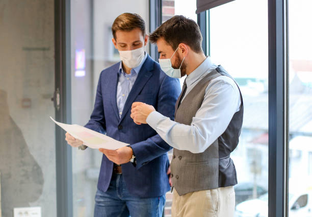 istock Two white collar worker with their protective masks consulting each other in front of window, near their office. 1292791513