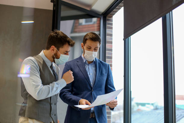 istock Two young experts having serious talk about printed charts in front of them. They have p[protective masks on and they are standing in front of the opened window. 1292791527