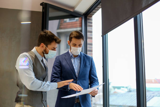 istock Two responsible workers standing next to opened window with protective masks on and talking about printed charts. their protective masks on 1292791531
