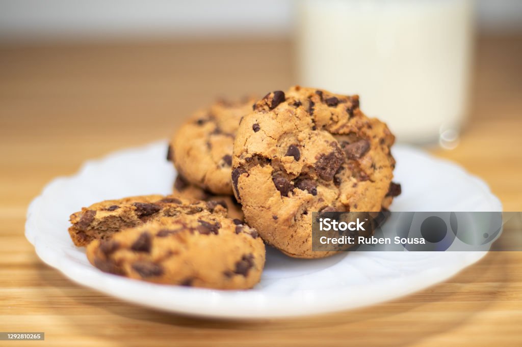 Chocolate chip cookies with a shallow dept of field, with glass of milk in the background Chocolate chip cookies with a shallow dept of field, with glass of milk in the background, on a wooden table. American Culture Stock Photo
