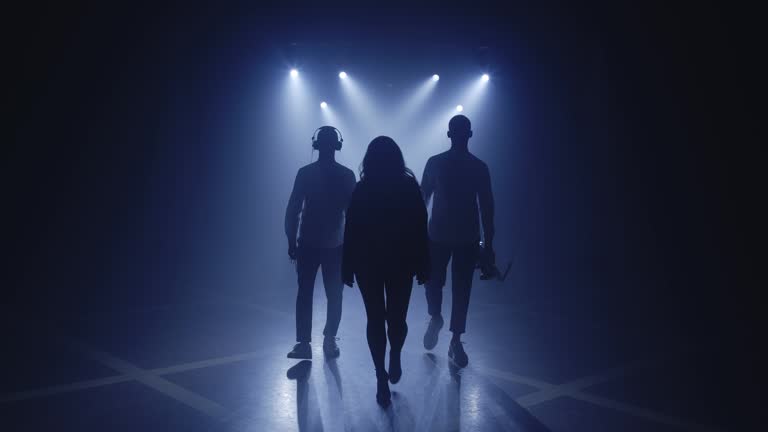 Backlit silhouettes of singer vocalist girl, saxophonist sax, dj man with headphones walking forward