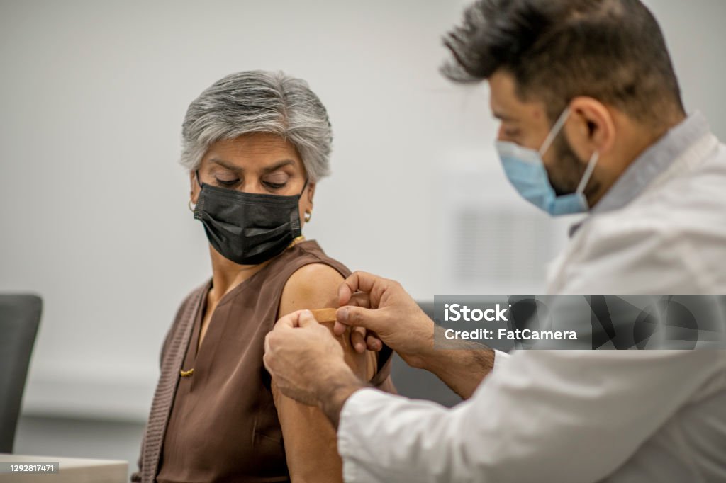 Putting band aid on after vaccination A male doctor puts a band aid on a senior woman's arm after he administered the COVID-19 vaccine injection. They are both wearing a protective face mask to protect themselves from the transfer of germs. Vaccination Stock Photo