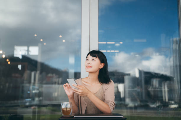 young asian woman looking up and planning on her project while using her smartphone and laptop outdoors in an urban park - looking up young women outdoors laptop imagens e fotografias de stock