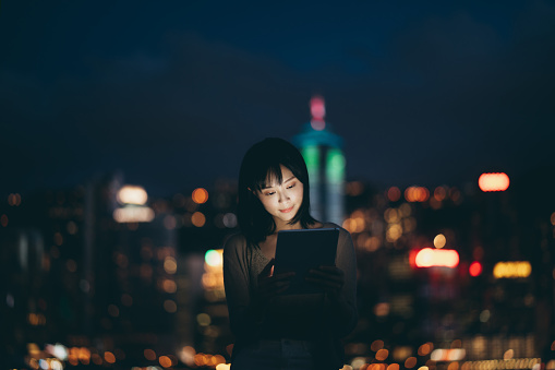 Young Asian woman using her laptop in the city, against illuminated city scene with highrise corporate skyscrapers at dusk