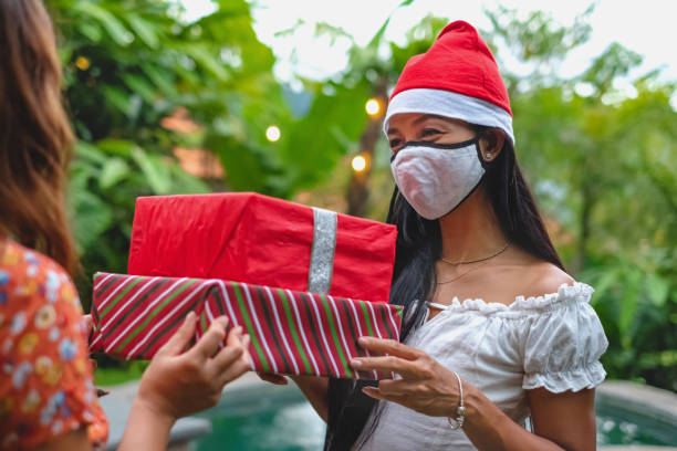 Indonesian woman receiving christmas gift from friend Close up shot of Indonesian woman with face mask and santa hat, receiving gifts from her friend outside her holiday villa during Covid 19 pandemic vacation rental mask stock pictures, royalty-free photos & images
