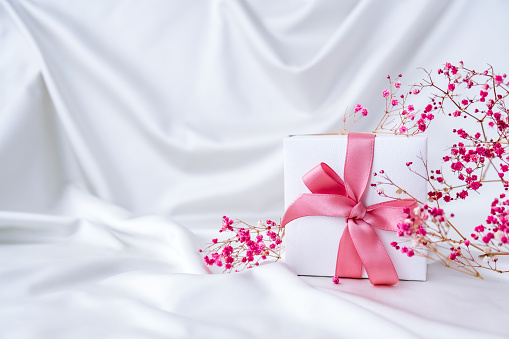 White present box with pink ribbon and small pink flowers on white silk fabric background. Greeting card for holidays. Copy space. Weeding Birthday Valentines Mothers Womans Day