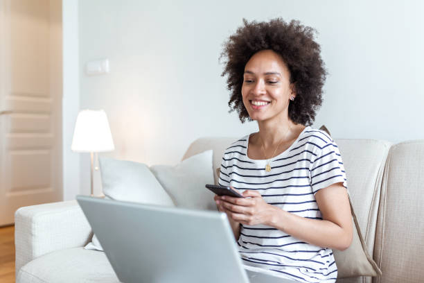 istock Happy young black woman using mobile phone while sitting a couch at home with laptop computer. Smiling woman sitting on sofa relaxing while browsing online shopping website. 1292851668