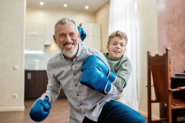 Portrait of a cheerful grandpa and little boy grandson wearing blue boxing gloves having fun and smiling at camera while having boxing training Portrait of a cheerful grandpa and little boy grandson wearing blue boxing gloves having fun and smiling at camera while having boxing training. Physical activity and sport for kids at home old man boxing stock pictures, royalty-free photos & images