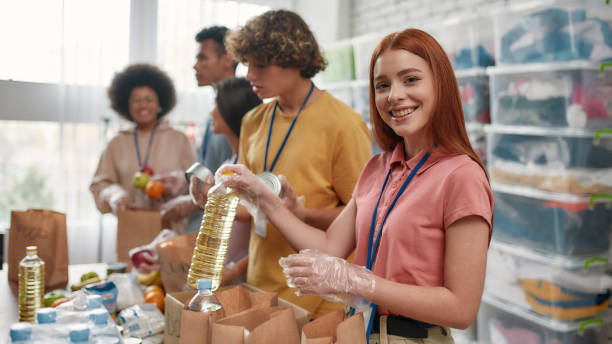 Young female volunteer smiling at camera while packing food and drinks donation into paper bags and box, Small group of people working in charitable foundation Young female volunteer smiling at camera while packing food and drinks donation into paper bags, Small group of people working in charitable foundation, Selective focus on girl, Web Banner doing a favor stock pictures, royalty-free photos & images