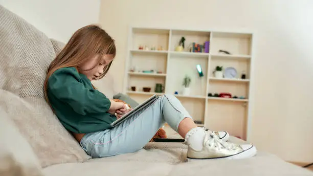 A cute little girl playing videogames on a tablet sitting on a sofa having bookshelves on a background of a spacy room staying alone at home. Children and modern technologies