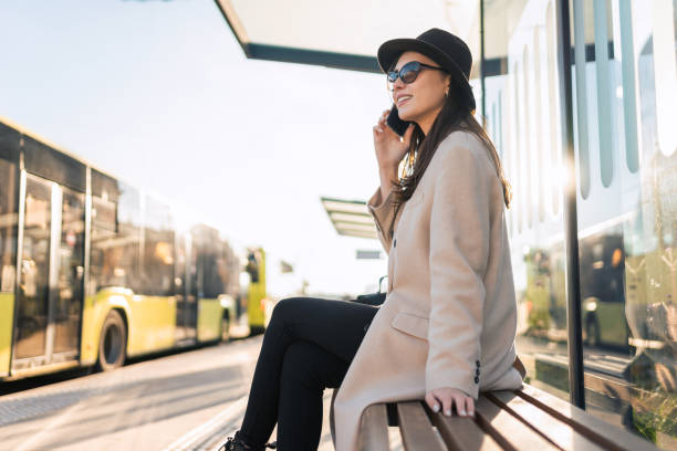 casual woman in glasses sitting at the bus stop and talking on the mobile phone. passenger waiting for city bus - color image bus discussion expertise imagens e fotografias de stock