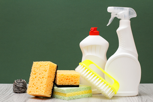 Plastic bottles of dishwashing liquid, glass and tile cleaner, brush and sponges on laminate flooring and green background. Washing and cleaning concept.
