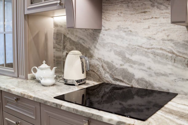 beautiful closeup of custom designed kitchen, with marble looking quartz countertop and backsplash. cream electric kettle with porcelain tea accessories on the marble countertop next to ceramic hob - quartz imagens e fotografias de stock